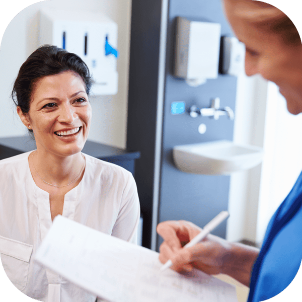 A female patient smiling and engaging with a healthcare provider who is holding a clipboard, in a medical examination room setting.