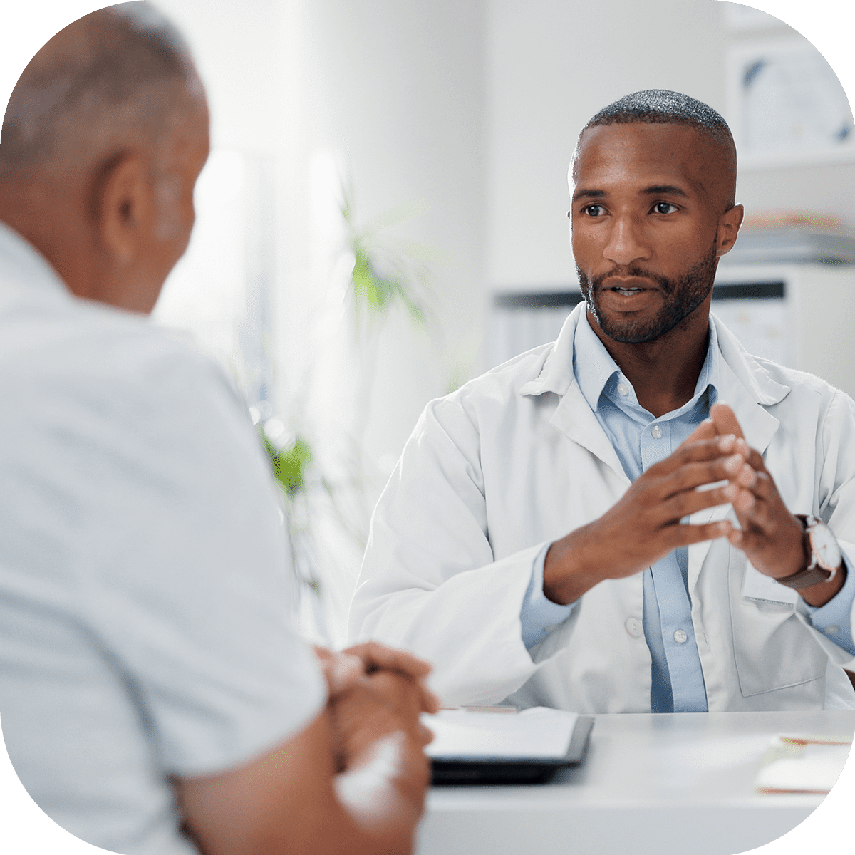 A male doctor in a white coat sits at a desk, attentively speaking and gesturing with his hands, while a male patient listens across from him in a medical office setting.